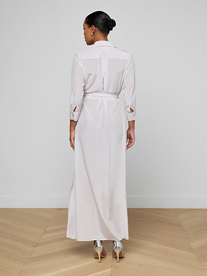 A woman with her hair in a neat bun stands on a wooden floor against a plain light background, wearing the Cameron Silk Shirt Dress—an ankle-length, long-sleeved silk crepe de chine dress with a belt—and metallic high-heeled sandals.