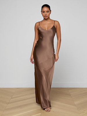 A woman stands indoors on a wooden floor wearing the Serita Silk Gown, a long brown slip dress with thin straps and open-toe heels. The plain, light background highlights this elegant choice for a wedding guest dress.