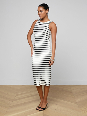 A woman poses indoors against a light wall and wooden floor, wearing the Nura Racerback Midi Dress—sleeveless with horizontal black stripes—and black open-toe heels.