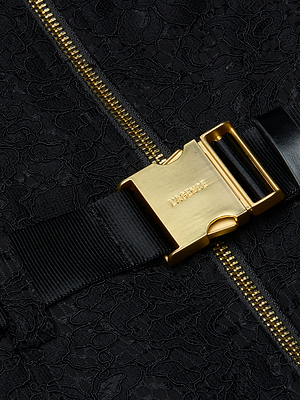 Close-up of a gold LACOSTE-engraved buckle clasp on a black woven strap, set against the textured black floral lace fabric of the Kylo Lace Shirt Dress with a visible gold zipper.
