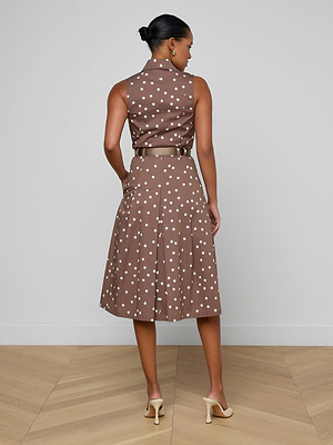 Woman wearing a brown polka dot dress standing on a wooden floor with a light gray wall background