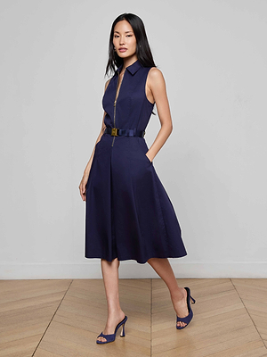 A woman with long black hair models the Kylo Poplin Shirt Dress—sleeveless, collared, navy blue with a matching belt—paired with high heels, standing on a wooden floor against a plain light gray wall.
