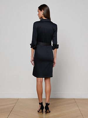 A woman faces away from the camera wearing the knee-length Karin Poplin Shirt Dress by L'AGENCE with rolled sleeves and black high-heeled sandals, standing on a wooden floor against a plain light wall.