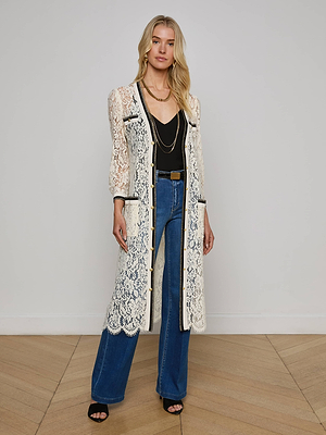 A woman poses against a plain wall in the Whisper Lace Cardigan Dress over a black top and blue wide-leg jeans, accessorized with layered necklaces and black sandals.