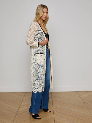 A woman stands sideways in the Whisper Lace Cardigan Dress, a long cream lace coat with black trim and scalloped edges, paired with blue jeans and black heels, against a plain white wall and wooden floor.