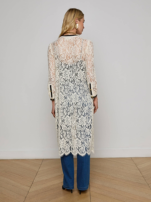 A person with long blond hair faces a white wall, wearing the Whisper Lace Cardigan Dress—a sheer, white lace piece with scalloped edges, long sleeves, and a below-the-knee length—over blue jeans and black shoes.