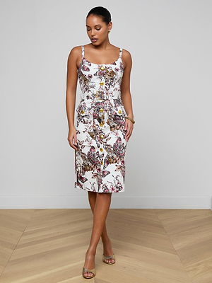 A woman stands indoors on a light wood floor, wearing the Ida Dress—a tailored stretch cotton piece with a vibrant butterfly jungle print—and open-toed heels. She looks down, one leg crossed in front of the other.