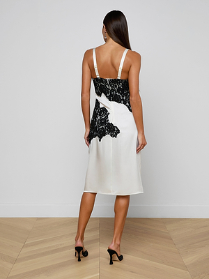 A woman with long, straight brown hair faces a white wall, wearing the Fenna Silk Lace Dress—an ivory silk midi slip with black lace and a sculpted bustier—paired with black heeled sandals on a light wood chevron floor.