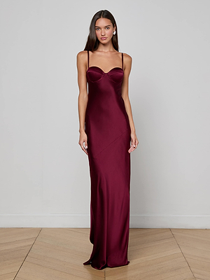 A woman with long, straight hair stands by a light wall wearing the L'AGENCE Sayla Silk Gown—an event-ready, deep burgundy dress featuring thin straps and a fitted bodice. She poses with her arms relaxed by her sides.