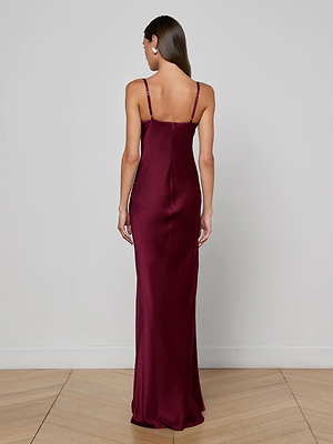 A woman with straight brown hair stands with her back to the camera in a minimalist room, wearing the L'AGENCE Sayla Silk Gown—a sleeveless, floor-length maroon dress with thin straps.