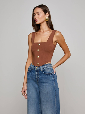 A woman with long brown hair wears the Saga Top—a chocolate brown, corset-inspired piece with gold front buttons—paired with high-waisted blue wide-leg jeans as she poses with one hand on her hip against a plain background.