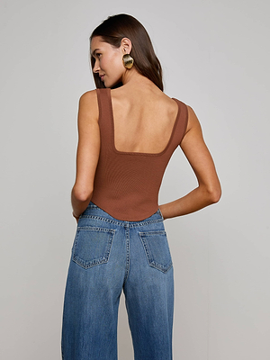 A woman with long brown hair wears the chocolate brown Saga Top, featuring a square neck and corset-inspired design, paired with blue jeans and large gold hoop earrings. She faces away, looking to the side against a plain background.