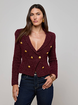 A woman with long brown hair models the L'AGENCE Bethany Knit Blazer in textured burgundy with gold buttons and dark blue jeans, accessorized with hoop earrings and gold bracelets, posed before a light gray backdrop.