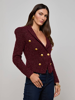 A woman with long brown hair models the Bethany Knit Blazer by L'AGENCE, a figure-flattering burgundy piece with gold buttons. She pairs it with dark jeans, gold earrings, and bracelets against a plain light background.