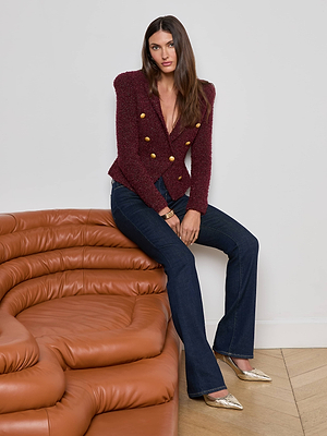 A woman with long brown hair sits on a tan leather sofa arm, wearing L'AGENCE's Bethany Knit Blazer in maroon with gold buttons, dark blue flared jeans, and gold pointed shoes, posing against a plain white wall.