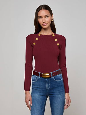 A woman with long, straight brown hair and gold earrings faces forward, wearing the maroon Gretchen Pullover by L’AGENCE with gold lion head buttons on the shoulders, blue jeans, and a maroon belt with a gold buckle.