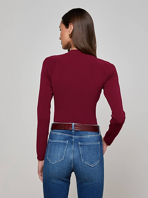 A woman with straight brown hair faces away from the camera, wearing the L'AGENCE Gretchen Pullover and high-waisted blue jeans with a textured brown belt. The background is plain light gray.