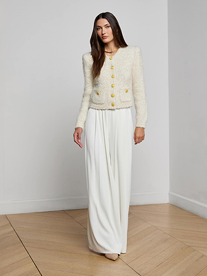 A woman with long brown hair stands in a minimal, light-colored room wearing the Una Knit Cardigan with gold buttons, wide-leg white pants, and neutral shoes.