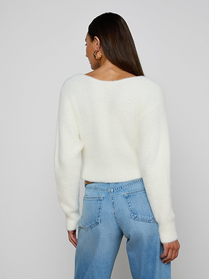 A woman with long brown hair is shown from the back, wearing the oversized ivory Antonella Crossover Sweater with light blue jeans and gold hoop earrings, standing against a plain light background.