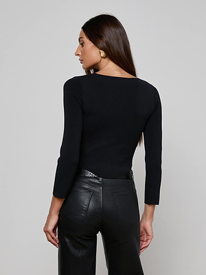 Woman with long brown hair wears the L'AGENCE Sasha Top, a corset-inspired sculpting stretch knit, paired with high-waisted black leather pants. Gold earrings accent her look as she poses against a light background, head turned to the side.