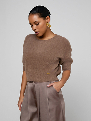 A woman stands against a plain backdrop with one hand in her pocket, wearing the Guinevere Cropped Pullover—a brown ultra-soft knit crewneck sweater—paired with silky high-waisted taupe pants, gold hoop earrings, and slicked-back hair.