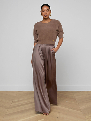 A woman stands against a plain background wearing the Guinevere Cropped Pullover—a brown ultra-soft knit crewneck sweater—with high-waisted, wide-leg taupe satin pants and one hand in her pocket. The floor is light wood.
