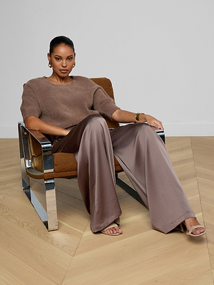 A woman sits confidently in a modern chair, wearing the Guinevere Cropped Pullover—a brown ultra-soft knit crewneck sweater with a cropped fit—paired with wide-leg satin trousers, open-toe heels, gold hoop earrings, and a bracelet in a minimalist room.