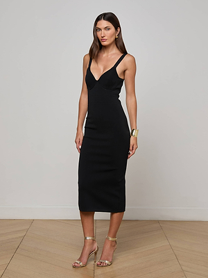 A woman stands indoors against a plain wall, wearing the Jimena Knit Dress with a sculpting silhouette, gold high-heeled sandals, and a chunky gold bracelet, her hair down and parted in the middle.