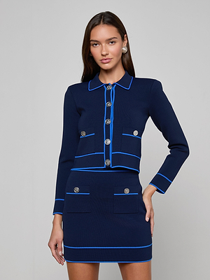 A woman with long brown hair and pearl earrings models the Tala Cardigan, a navy blue stretch knit suit with light blue trim, silver buttons, two skirt pockets, and two chest pockets on the jacket against a plain background.