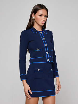 A woman with long brown hair wears the Tala Cardigan, a navy blue power knit skirt suit with light blue trim, silver buttons, and front pockets. She stands against a plain background and accessorizes with silver earrings.