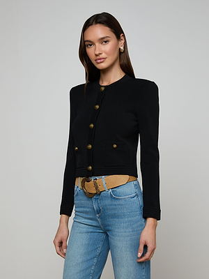 A woman with long dark hair wears the Patsy Cropped Cardigan in black with signature buttons, light blue jeans, a tan belt, and gold earrings, standing against a plain light gray background.