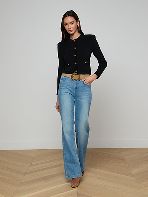 A woman stands indoors wearing the Patsy Cropped Cardigan, light blue high-waisted jeans, a tan belt, and nude heels. She poses with one hand in her pocket against a simple, light-toned wall and wooden floor.