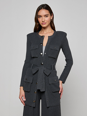 A woman with long brown hair models the Lore Cardigan, a dark grey knit sweater jacket featuring a tie belt, large buttons, and multiple front pockets, against a plain light grey background.