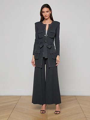 A woman stands on a wooden floor by a light gray wall, wearing the dark gray Lore Cardigan with front pockets and a tie belt, matching wide-leg trousers, and black open-toe heels.
