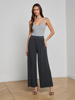 A woman stands indoors on a wooden floor, wearing a fitted grey tank top, the charcoal Milly Knit Pant with a pull-on high-waisted wide-leg fit, and heeled sandals, one hand on her hip and a neutral expression.
