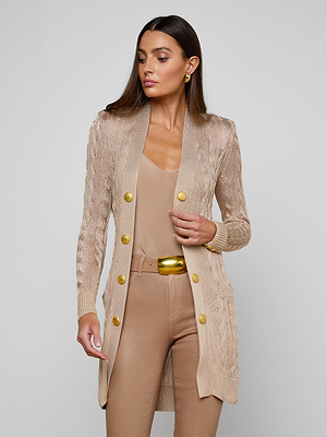 A woman with long, straight brown hair wears the Penelope Cabled Cardigan in camel, styled with a matching beige top and pants plus a gold belt. She stands against a light background, looking slightly to her left.