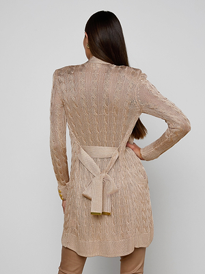 A woman with long brown hair faces away, wearing the Penelope Cabled Cardigan in camel with a tied waist belt and sleeve logo buttons, paired with matching brown pants and one hand on her hip.