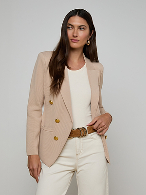 A woman with long brown hair wears the Amelia Knit Blazer—a structured beige style with gold buttons—over a white top and light jeans, accessorized with a brown belt and gold hoop earrings, against a plain light background.