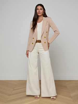 A woman poses indoors against a plain wall and wooden floor, wearing the Amelia Knit Blazer in tan with gold buttons, a white top, cream high-waisted wide-leg pants, a brown belt, and open-toe shoes.