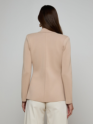 A woman with long brown hair is seen from the back, wearing the Amelia Knit Blazer in beige with gold buttons and white pants, standing against a plain light gray background.
