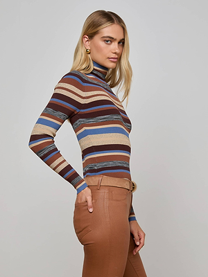 A woman with long blonde hair wears the L'AGENCE Olene Turtleneck, a ribbed knit sweater with lurex stripes in brown, blue, and beige, paired with high-waisted brown pants. She stands sideways, gazing neutrally at the camera.