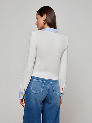 A woman stands with her back to the camera, wearing the April Poplin Collar Pullover—a white sweater with a striped poplin collar and blue striped cuffs—paired with blue jeans and gold hoop earrings against a plain, light background.
