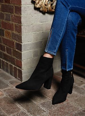 A person wearing blue zippered skinny jeans and black pointed-toe heeled ankle boots sits with legs crossed on a brick floor near a cream-colored brick wall.
