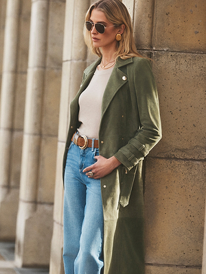 A woman in sunglasses, a light beige top, high-waisted blue jeans, and a Celina Suede Trench Coat stands by a stone column with one hand in her pocket—a perfect choice for a timeless wardrobe.