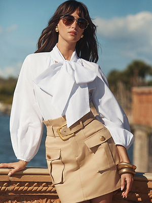A woman in sunglasses and the Deon Tie-Neck Blouse with blouson sleeves, paired with a beige belted mini skirt with large pockets, stands outdoors resting her arm on a stone balustrade under a sunny sky.