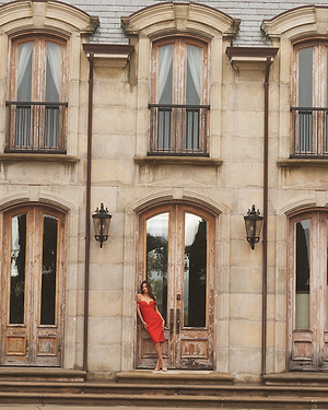 A woman in a red dress stands on the steps in front of a large, elegant stone building with tall, arched wooden doors and windows.
