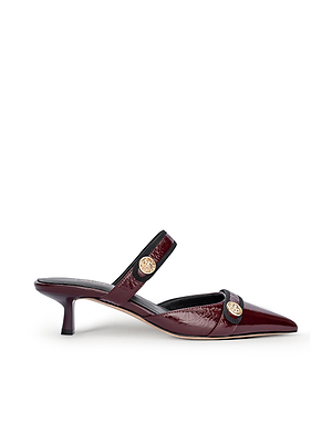 The Gilliane Heeled Mule by L'AGENCE features wine patent leather, a pointed toe, low kitten heel, and two thin straps with gold lion-embossed buttons—offering a chic blend of vintage and contemporary style.