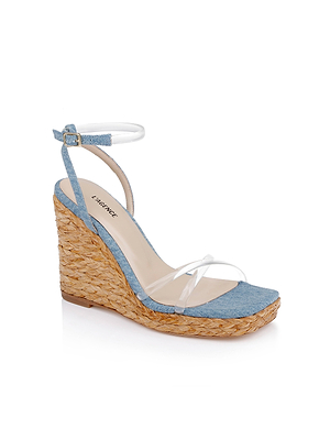 The Malone Wedge Espadrille features a woven raffia wedge heel, light blue denim toe strap, clear toe strap, and a white ankle strap with buckle. Displayed on a white background.