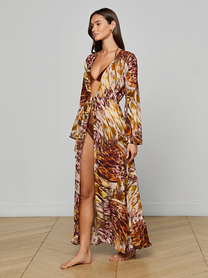 A woman stands barefoot on a wooden floor, wearing the Janus Cover-Up Robe with a feather print, deep neckline, high slit, and a brown bikini underneath. She gazes to the side against a plain light gray wall.