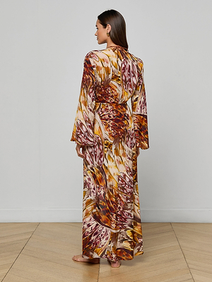 A woman stands barefoot on a wooden floor, facing away. She wears the Janus Cover-Up Robe, a long-sleeved, floor-length piece featuring an autumn-inspired abstract pattern in red, brown, yellow, and cream.